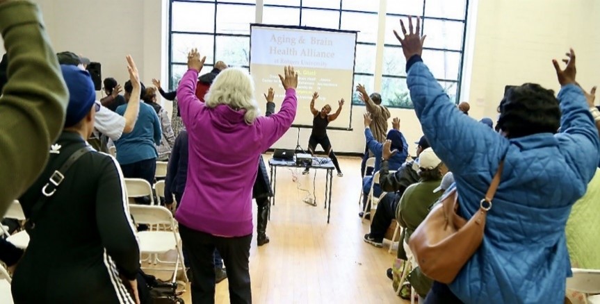 Young and older adults performing overhead stretches with an instructor in a large community room.